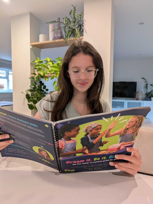 A young girl wearing glasses sits at a table reading the Dream it, Be it, Biz-it! workbook from We Can Be, smiling and engaged in a bright, modern home setting.
